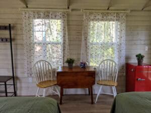 Dining area inside a Stonington cabin rental with a vintage wooden drop-leaf table centered between two windows with leafy-patterned sheer curtains, flanked by two white wooden chairs and a bright red retro mini-fridge.