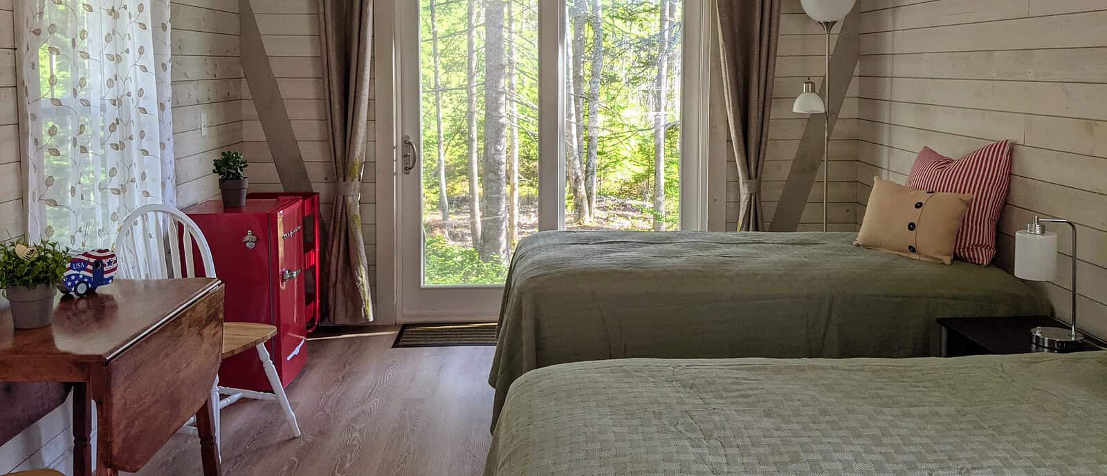 Interior of a cozy Maine cabin rental featuring two twin beds with green quilts, a rustic wooden drop-leaf table with white chairs, and a retro red Frigidaire mini-fridge against white wood-paneled walls under a vaulted ceiling with exposed beams. Glass sliding doors offer a scenic view of the surrounding Stonington woods.