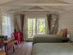 Interior of a cozy Maine cabin rental featuring two twin beds with green quilts, a rustic wooden drop-leaf table with white chairs, and a retro red Frigidaire mini-fridge against white wood-paneled walls under a vaulted ceiling with exposed beams. Glass sliding doors offer a scenic view of the surrounding Stonington woods.