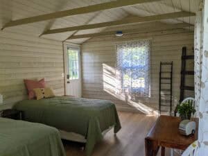 Sunlit interior of a Deer Isle camping cabin rental showing twin beds with green quilts, a wooden table, and sunlight streaming across white wood walls from a window with patterned sheer curtains near an entry door.