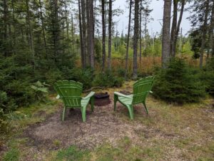 Outdoor campfire area at a rustic Maine cabin rental in Stonington, showing a stone-ringed fire pit surrounded by seating in a clearing of the pine woods.