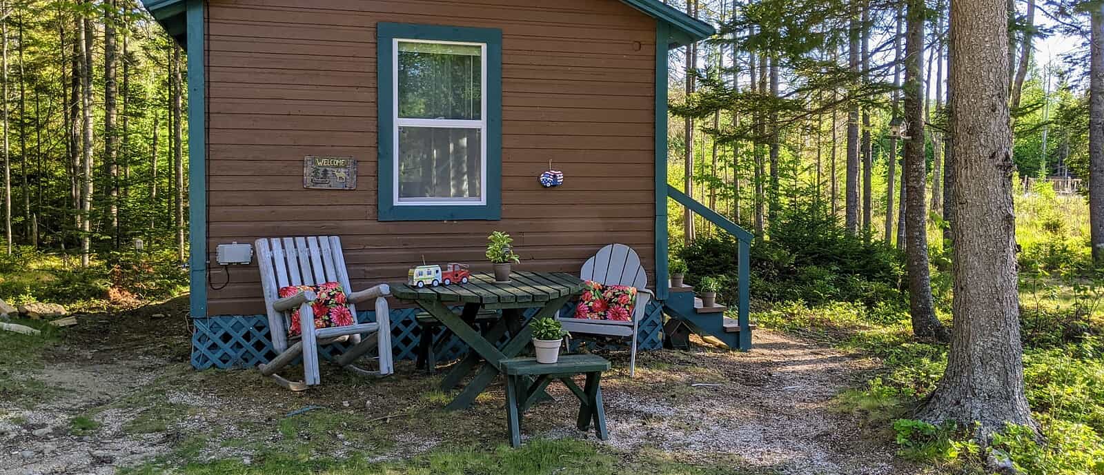 A charming brown vacation cabin rental at a Stonington Maine campground, surrounded by tall pine trees with an outdoor wooden table and chairs set up in the shaded yard for relaxing.