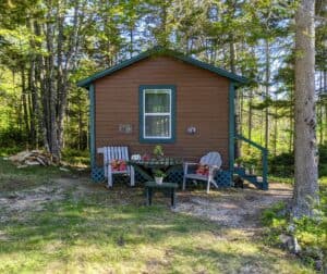 Exterior view of a brown rustic camping cabin with teal trim tucked into the Maine woods, featuring a private outdoor seating area with an Adirondack chair, a rocking chair, and a wooden picnic table.