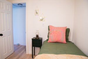 Bright bedroom inside Norma’s Cottage featuring a single bed with a green quilted bedspread, a red-and-white striped accent pillow, and a green display cabinet next to a sunny window