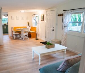 Spacious and sunlit living room of Norma's Cottage featuring a clean white coffee table, a teal sofa corner, a wooden dining nook, and bright windows looking out to the trees