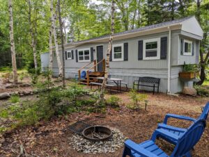 A peaceful outdoor seating area next to Norma's Cottage, showing blue Adirondack chairs positioned near a fire pit area surrounded by tall trees in the Maine woods