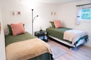 Bright bedroom inside Norma's Vacation Home featuring two twin beds with green quilted coverlets, red-and-white striped accent pillows, tan throw blankets, and a central black nightstand with a classic lamp