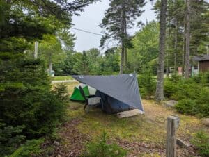 Tent campsite featuring a gray rain tarp canopy suspended over a tent and lawn chair under tall trees at Greenlaw's RV Park & Campground