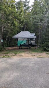 A pop-up camper trailer parked in a private, wooded campsite featuring a gray canopy and picnic table at Greenlaw's RV Park & Campground on Deer Isle near Stonington, Maine.